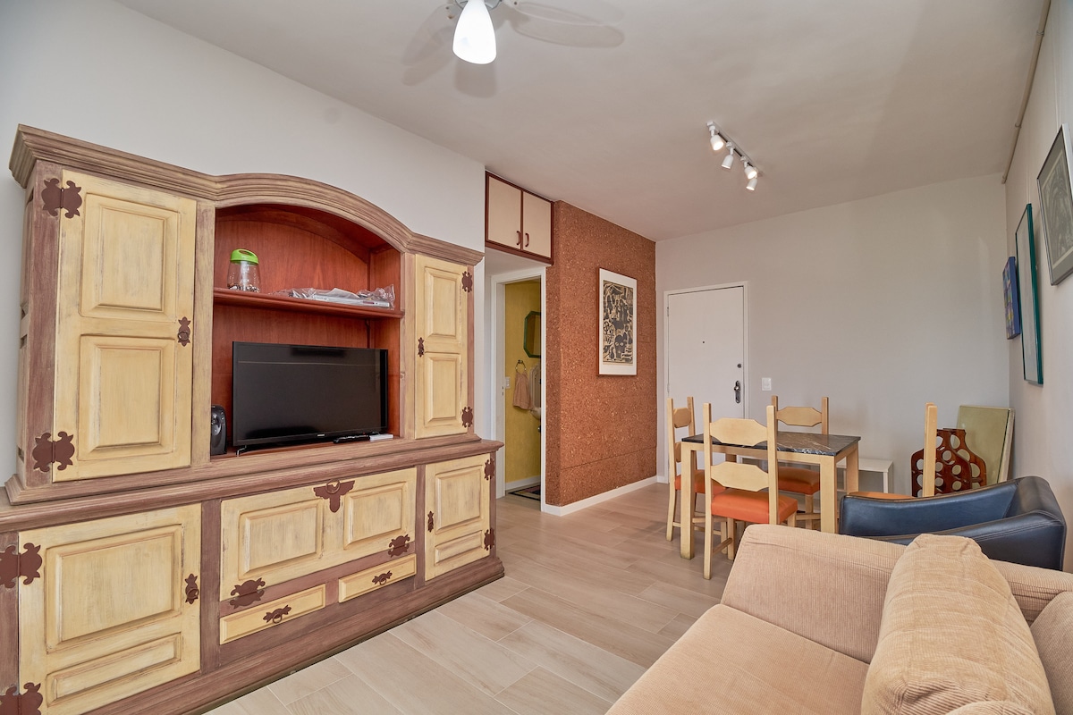 The living space features a cozy sofa, a wooden cabinet with a TV, and a dining area with multiple chairs and a table. A light-colored wall complements the warm tones of the furniture, while a ceiling fan provides air circulation.