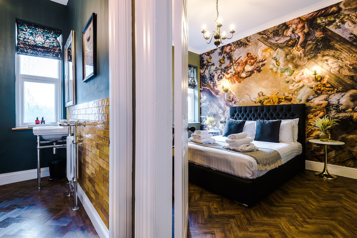 An elegantly appointed bedroom with a large bed adorned with neatly arranged linens. A striking mural enhances the wall above, while large windows allow natural light to brighten the space. An adjacent bathroom features stylish tiles and modern fixtures.