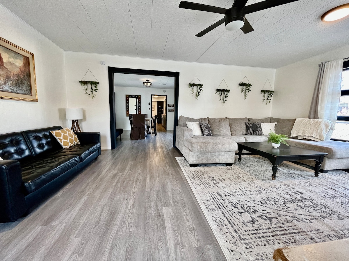 A spacious living room is highlighted by a large sectional sofa and a sleek black couch with patterned cushions. A coffee table sits centrally on a soft area rug. The room features light wood flooring and is adorned with wall plants and an artwork piece.