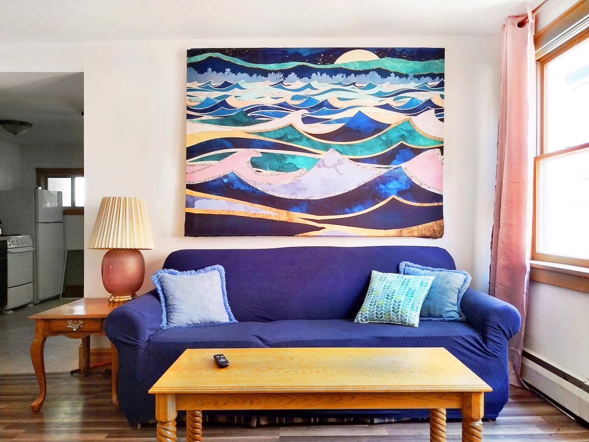 A cozy living area features a blue sofa with decorative pillows and a wooden coffee table. Above the sofa hangs a large, colorful painting of waves. Natural light enters through the windows, illuminating the warm tones of the space.
