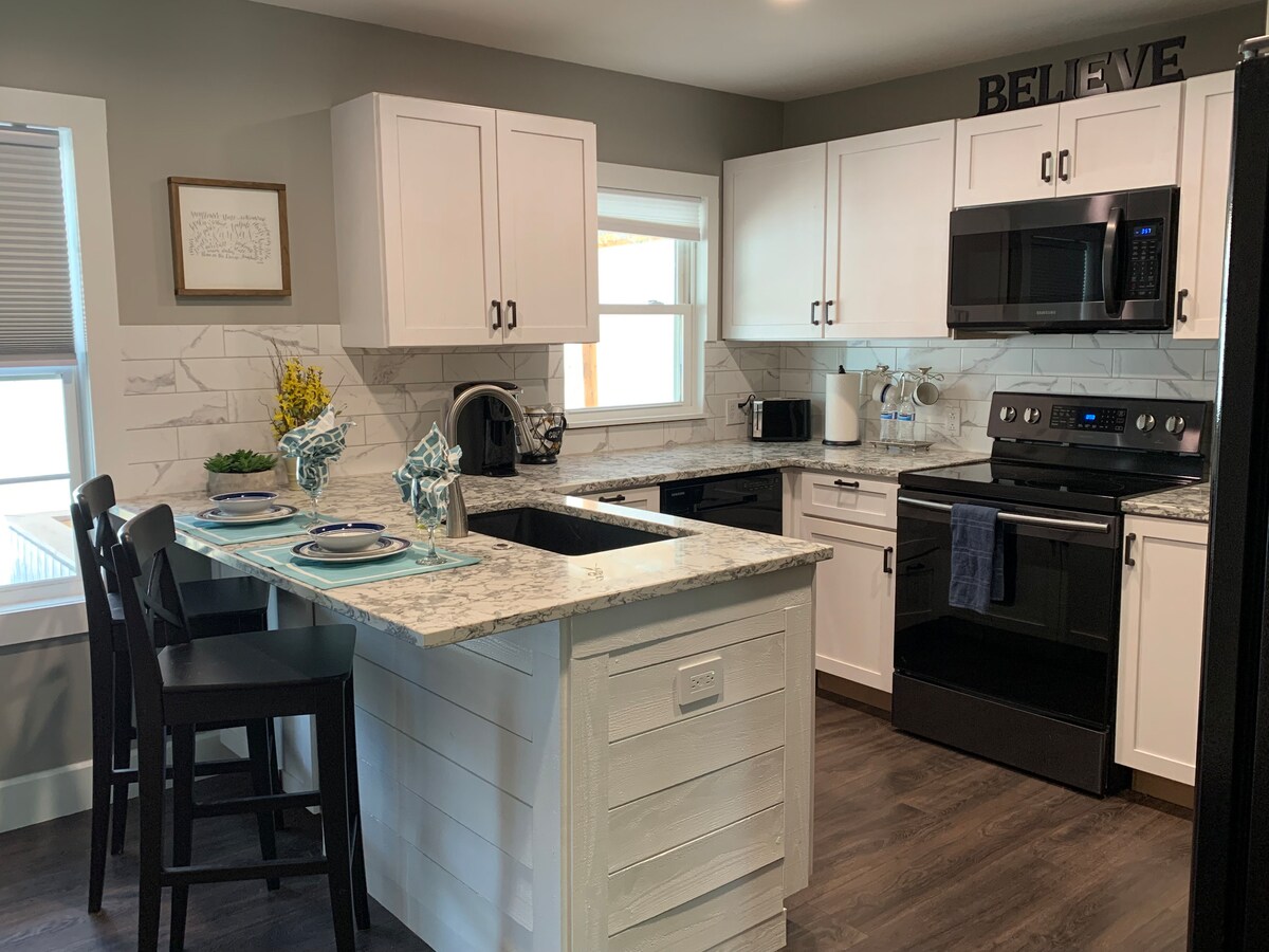 A modern kitchen is presented, featuring white cabinetry and a granite countertop. New appliances include a black microwave and refrigerator. A small dining area is visible, with a set table showcasing blue and white dishware. Natural light enters through a window above the sink.