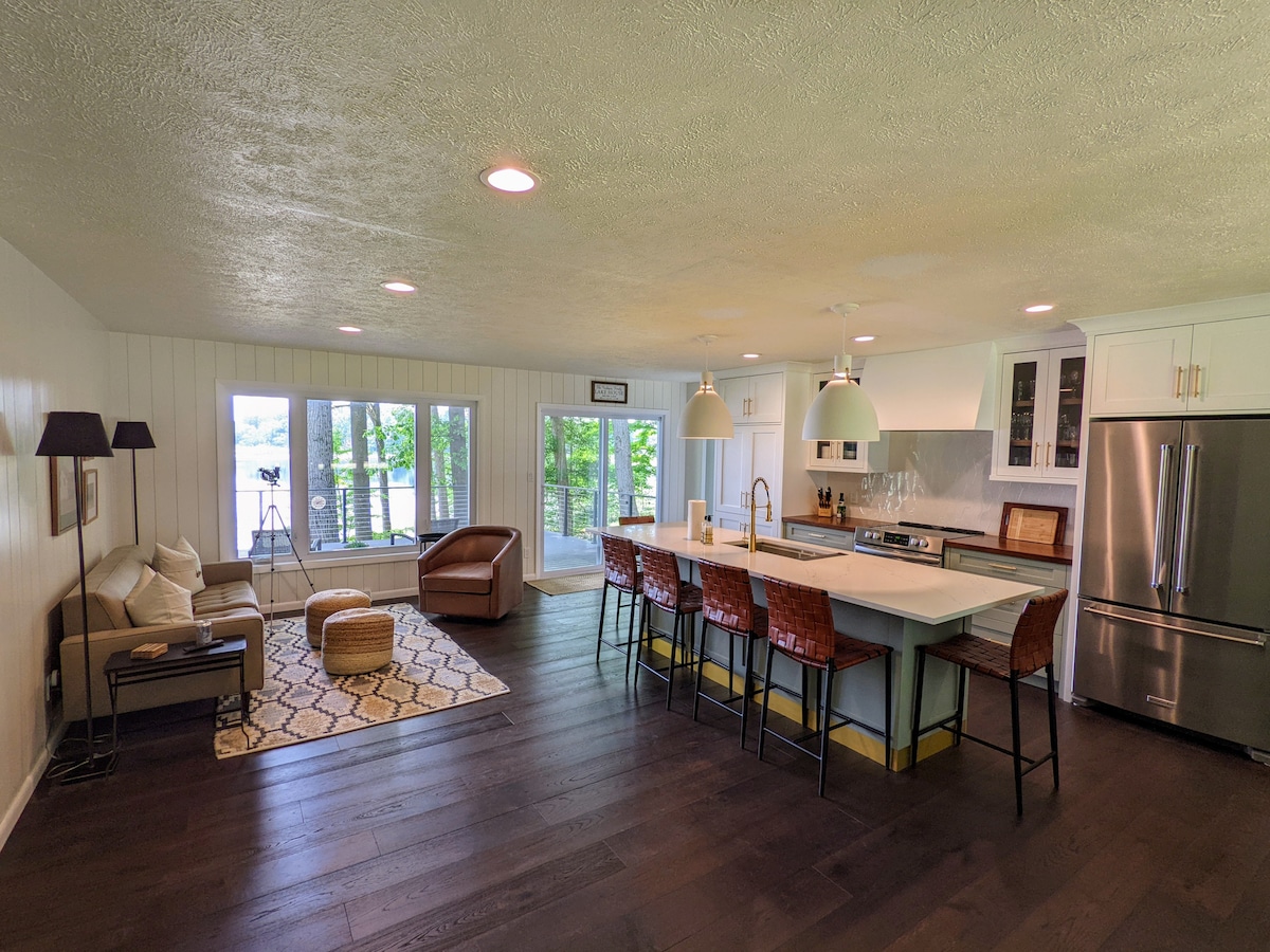 An open-concept living area showcases a modern kitchen with new appliances and a large island with seating. Natural light fills the space through expansive windows, which overlook the lake. A cozy seating arrangement includes a sofa and an accent chair, enhancing the inviting atmosphere.