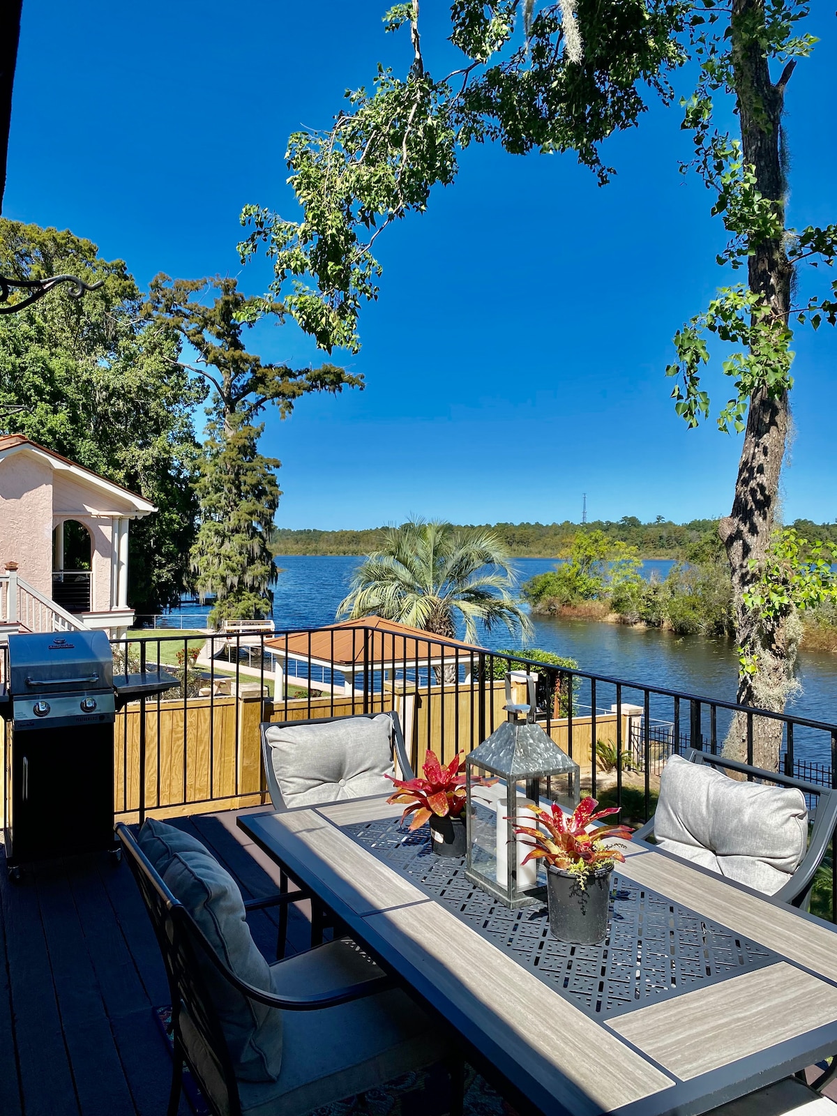 An outdoor deck overlooks the water, furnished with a table and chairs, set with decorative plants. A grill is positioned nearby, and trees provide shade, enhancing the serene lakeside environment.