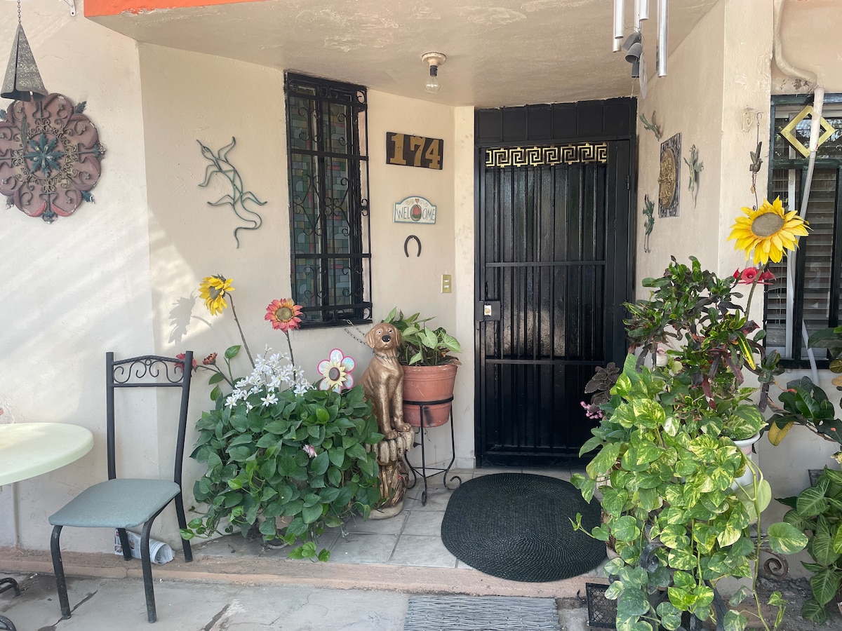 The entrance features a black door with a decorative design, framed by various potted plants, including vibrant flowers and greenery. A small round table with a chair is positioned nearby, providing a welcoming outdoor space enhanced by a joyful arrangement of decorative elements.