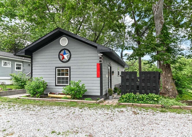 The exterior of the cozy casita is shown, featuring grey siding and a decorative star emblem. A black wooden fence borders the property, while lush greenery and a gravel driveway enhance the inviting atmosphere of the home.