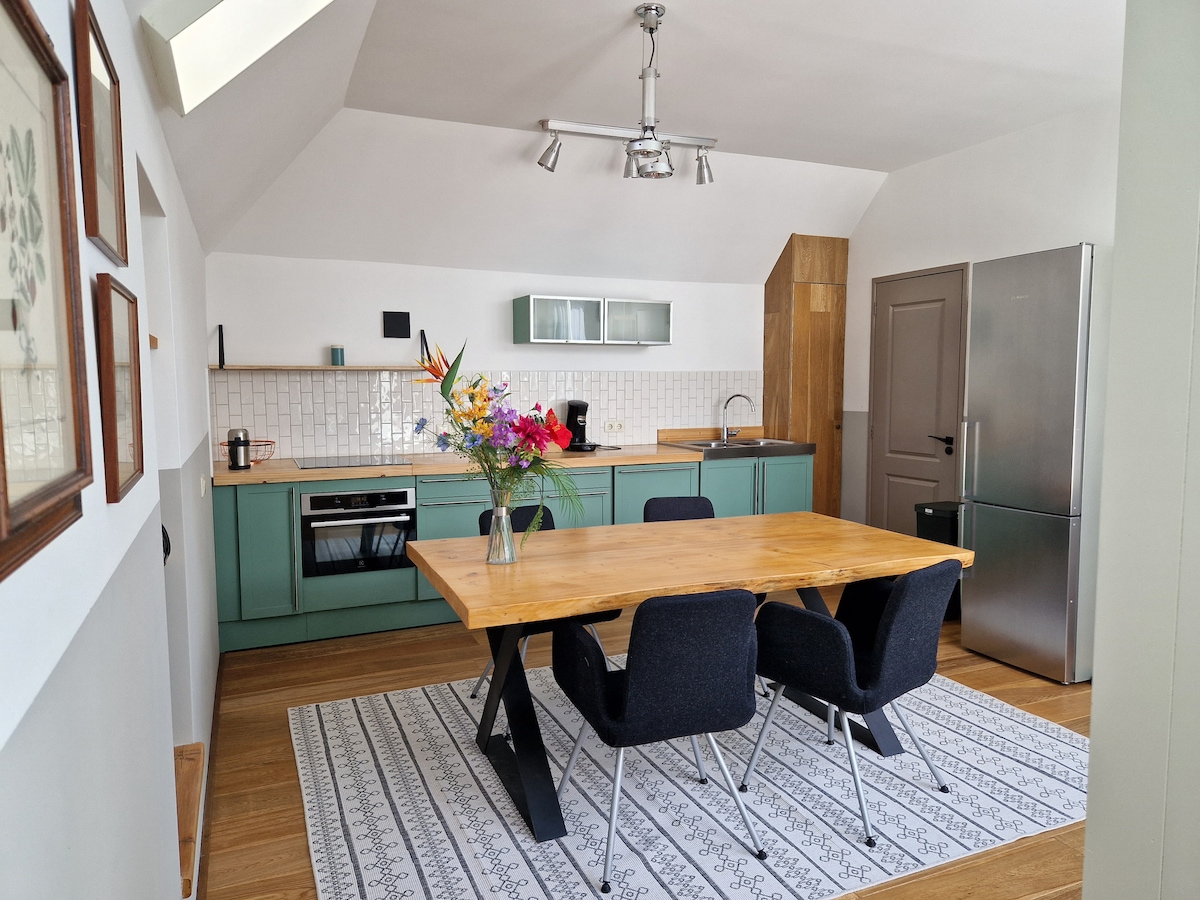 A modern kitchen and dining area feature a wooden table surrounded by black chairs. Light from a skylight highlights green cabinetry and stainless steel appliances. A floral arrangement decorates the table, and a patterned rug adds warmth to the wood flooring.