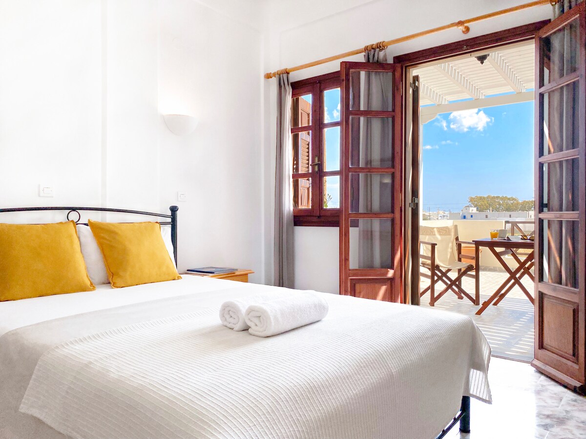 A comfortable sleeping area features a spacious bed with white linens and two yellow accent pillows. The room is brightened by natural light flowing through a set of wooden double doors that open to a private balcony, furnished with a table and chairs.