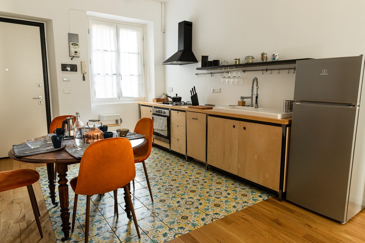 The kitchen area features wooden cabinetry and stainless steel appliances, including a refrigerator. A dining table with orange chairs is set for four, creating an inviting space. Colorful patterned tiles cover the floor, and natural light filters in through a large window with sheer curtains.