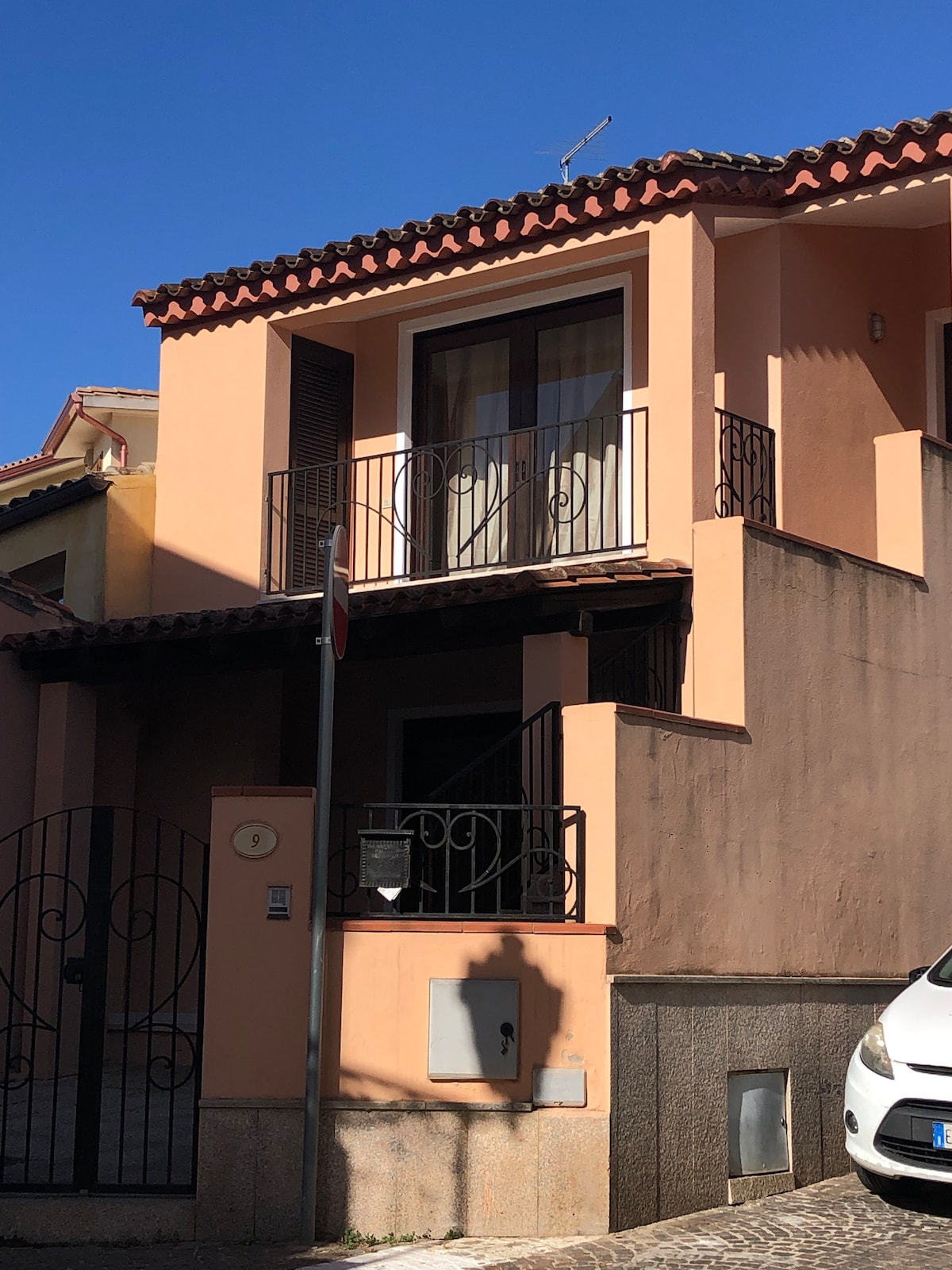 An exterior view of a two-story building with a pink façade and terracotta roof tiles is displayed. A small balcony on the upper floor features a railing. A white car is parked in front, and a decorative gate can be seen leading to the entrance.
