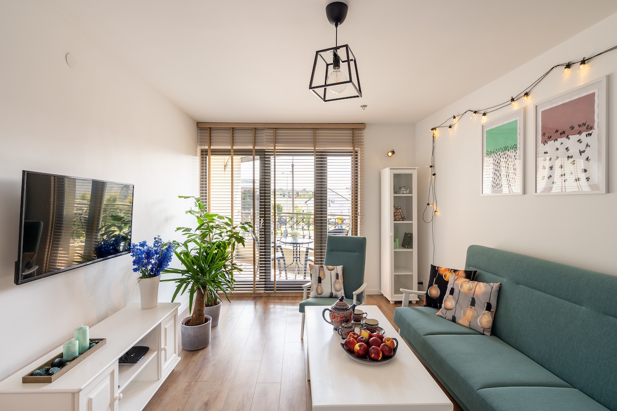 A bright living area features a green sofa adorned with patterned cushions, positioned across from a wall-mounted television. A white table holds a bowl of apples. Natural light filters through blinds, illuminating a potted plant and artworks on the walls.