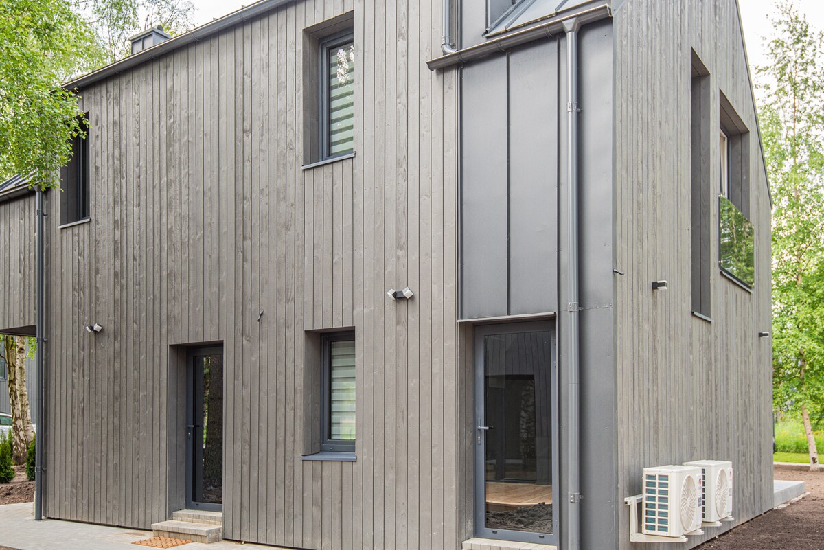The exterior of a modern two-story building is shown, featuring a sleek design with gray wood siding. Large windows are framed in dark metal, while a small path leads to the entrance. An air conditioning unit is located on the side of the structure.
