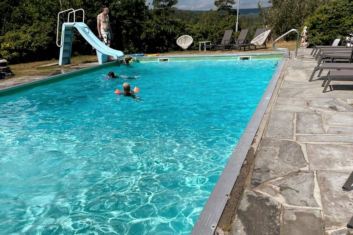 A large outdoor swimming pool is presented, featuring a sleek slide on one side. Several loungers are arranged beside the pool, and individuals can be seen enjoying the water. Surrounding greenery adds a natural ambiance to the area.