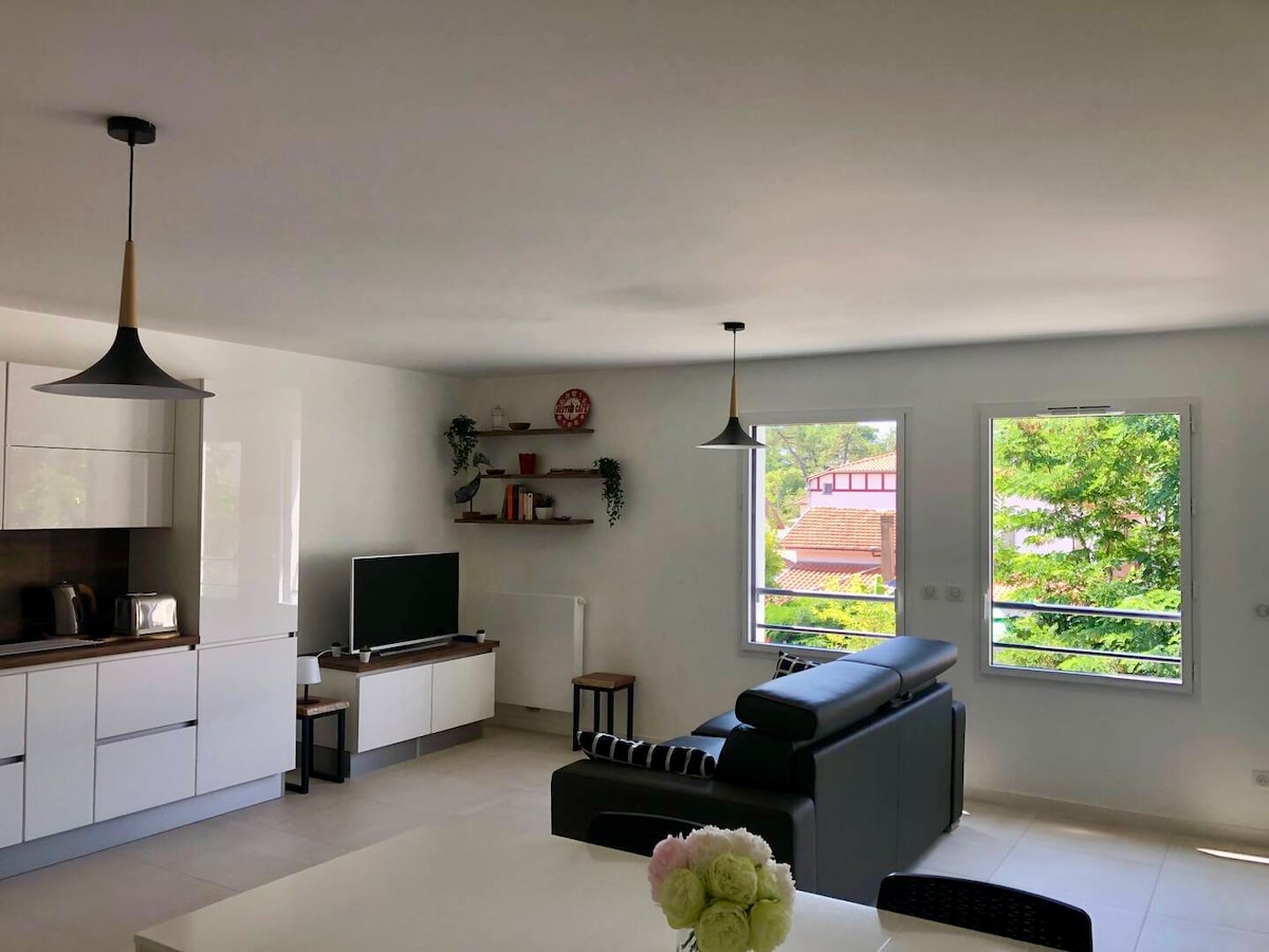 A spacious living area features large windows allowing abundant natural light. A modern sofa is positioned adjacent to a sleek television and minimalistic shelving. Two hanging light fixtures complement the bright, airy atmosphere, while a dining table is visible in the foreground.