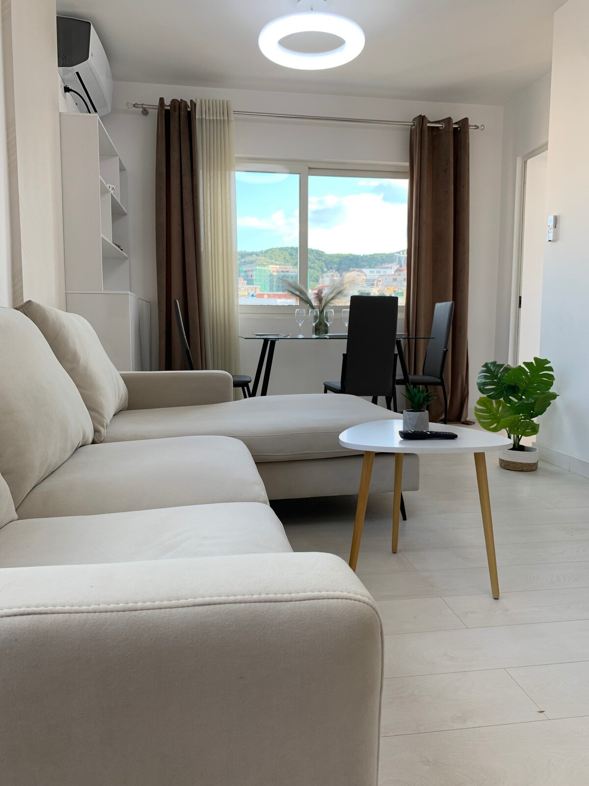 A light-filled living area is presented, featuring a spacious beige sofa and a round white coffee table. The space is adorned with potted plants and framed by large windows with sheer curtains, providing a view of the surrounding landscape. Nearby, a dining table and chairs are visible.