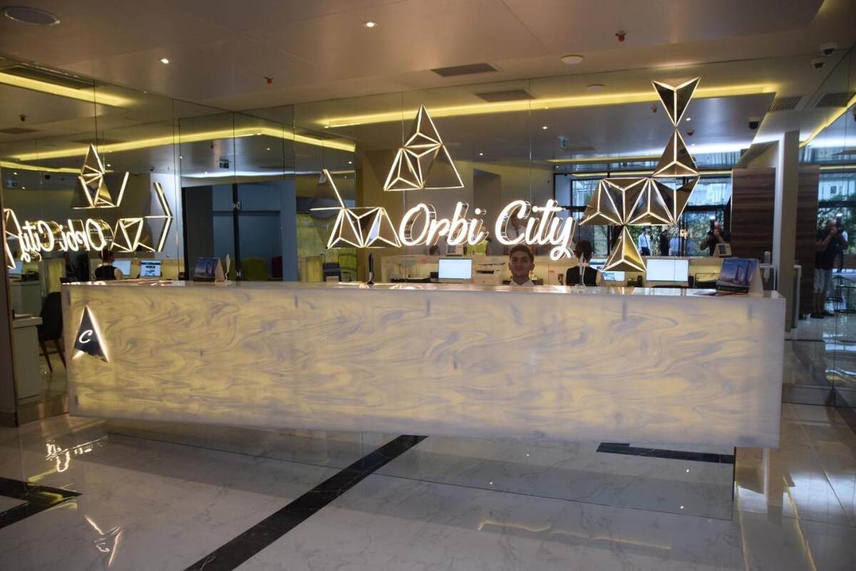 A modern reception area features a sleek, illuminated front desk adorned with geometric pendant lights. The backdrop showcases reflective surfaces and warm lighting, creating an inviting atmosphere. Monitors are positioned on the desk, and decorative elements enhance the contemporary design.