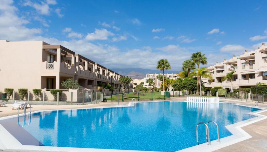 A spacious outdoor pool features clear blue water, surrounded by well-maintained gardens. Sun loungers are set up along the poolside, while palm trees provide shade. The two-story buildings are visible in the background, blending with the tranquil landscape and clear blue sky.