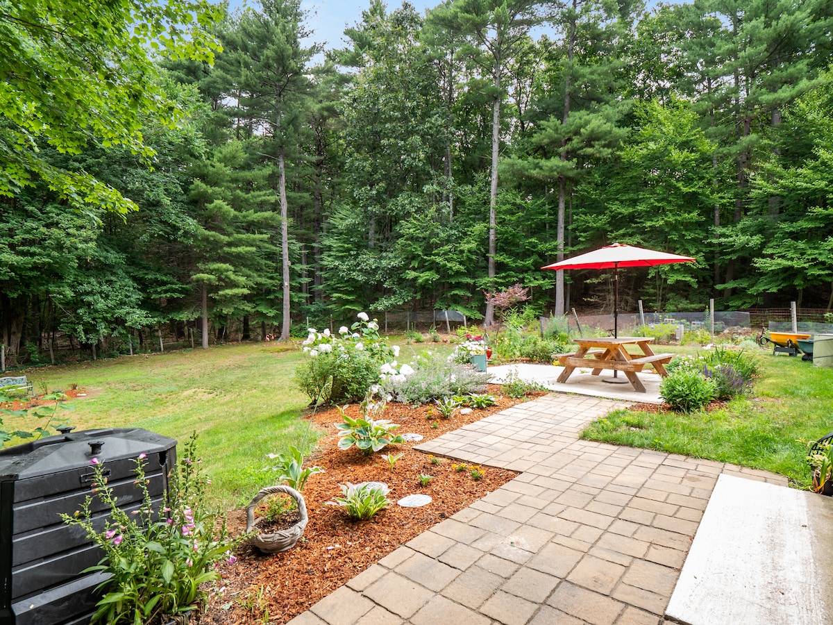 A spacious outdoor area features a picnic table shaded by a large red umbrella, surrounded by a well-maintained garden. Lush green trees form a natural backdrop, while pathways meander through flower beds and shrubs, creating an inviting space for relaxation.