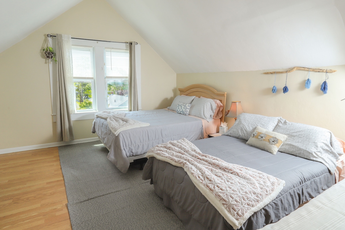 A bright bedroom features two beds, one queen and one full, positioned on a natural hardwood floor. A window allows ample natural light, complemented by soft fabric curtains. Bed linens in neutral tones create a calming ambiance, with decorative pillows adding subtle detail.