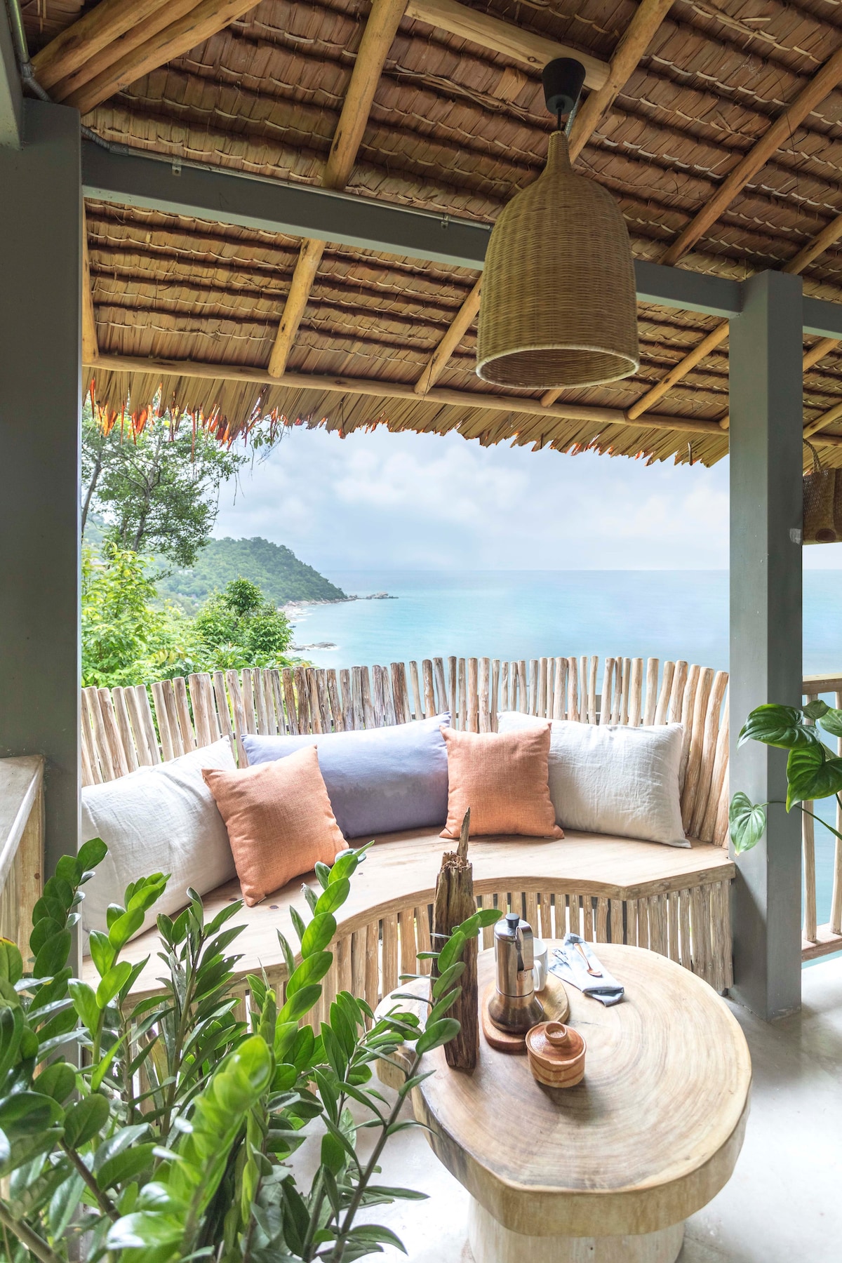 A comfortable seating area is featured on a balcony, surrounded by natural wooden fencing. The space includes plush cushions in warm tones and a round wooden coffee table. Lush greenery is visible, and the serene ocean view is framed by a thatched roof.