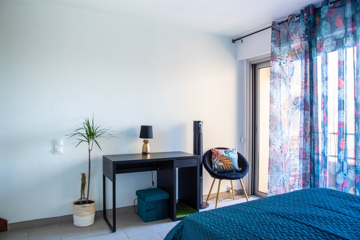 A bright bedroom includes a spacious bed with textured bedding and large windows covered by sheer floral curtains. A desk is positioned beside a stylish black chair, while a small potted plant and decorative items add touches of decor.
