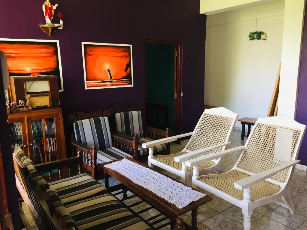 A comfortable sitting area is presented with several chairs featuring woven seats and a wooden table in the center. Colorful artwork adorns the walls, contrasting with a deep purple backdrop. Natural light brightens the space, enhancing its inviting atmosphere.