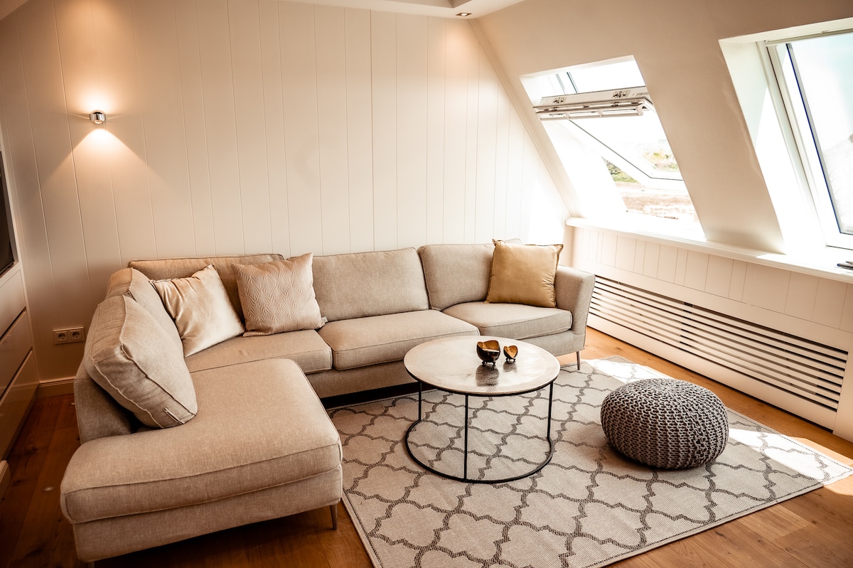 A light-filled living area features a spacious sectional sofa in soft beige, complemented by a textured gray pouf. A round coffee table rests on a patterned rug, while large windows invite natural light and offer a glimpse of the outdoor scenery.
