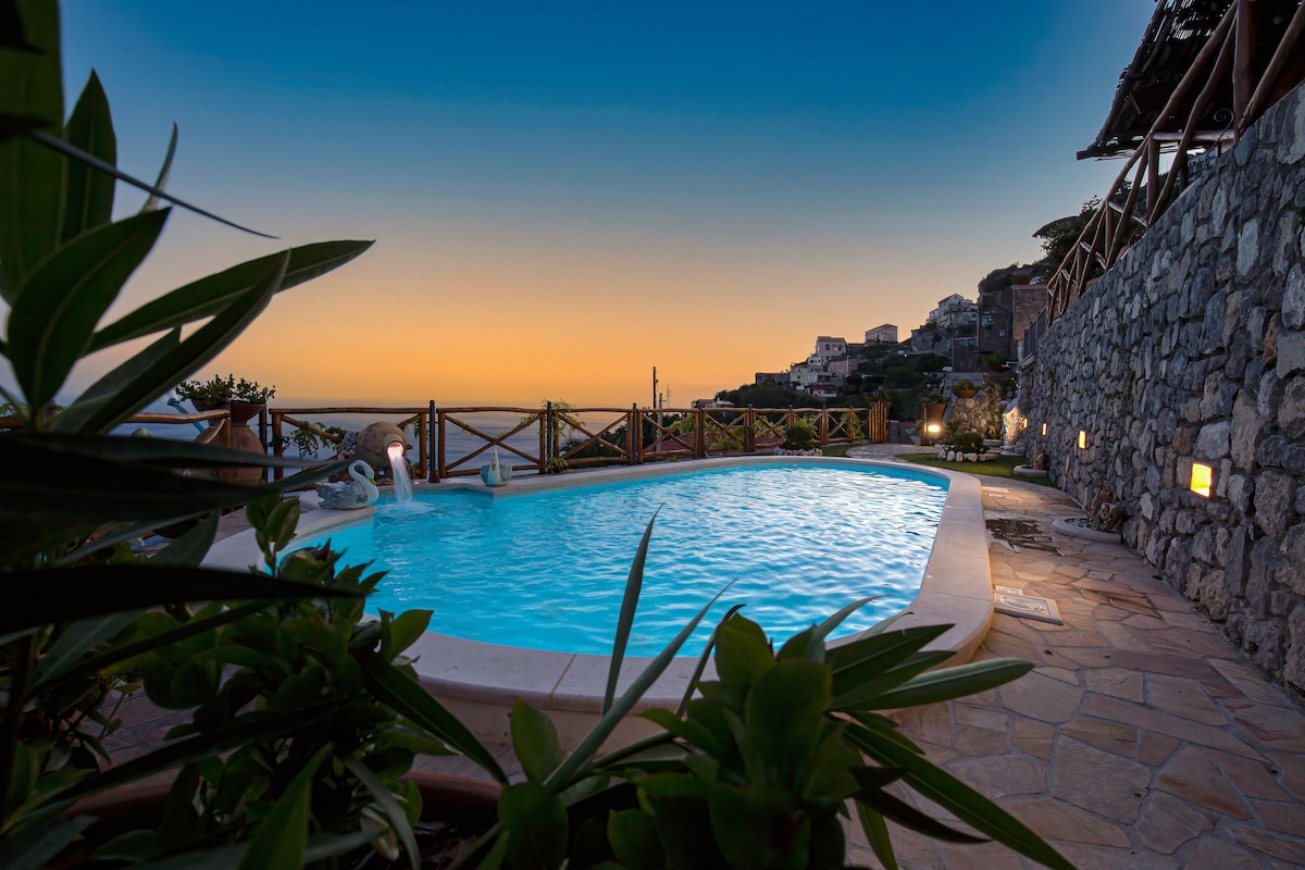 The swimming pool is bordered by natural stone, surrounded by lush greenery and gentle lighting. A serene water surface reflects the soft hues of the sunset, creating a peaceful atmosphere. The view extends towards the hillside village in the background, enhancing the tranquil setting.