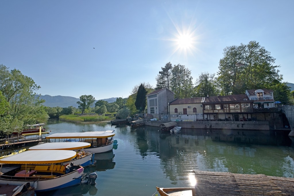 The peaceful scene captures a calm waterway, lined with greenery and buildings. Several boats are moored by the shore, while sunlight reflects off the water's surface. In the background, trees and a structure add to the tranquil atmosphere.