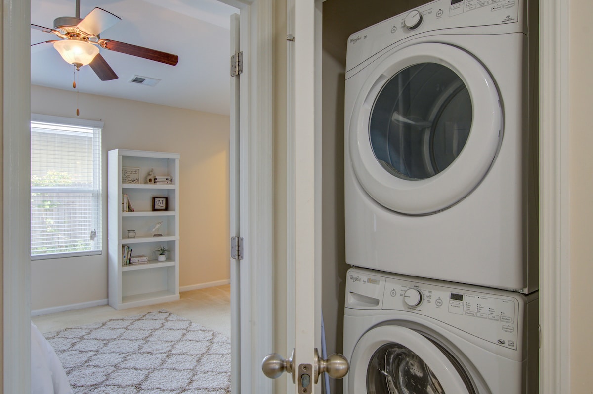 A laundry area is visible, featuring a stacked washer and dryer. A doorway leads to a well-lit bedroom with natural light filtering through a window, complemented by a white shelving unit displaying books and decorative items.