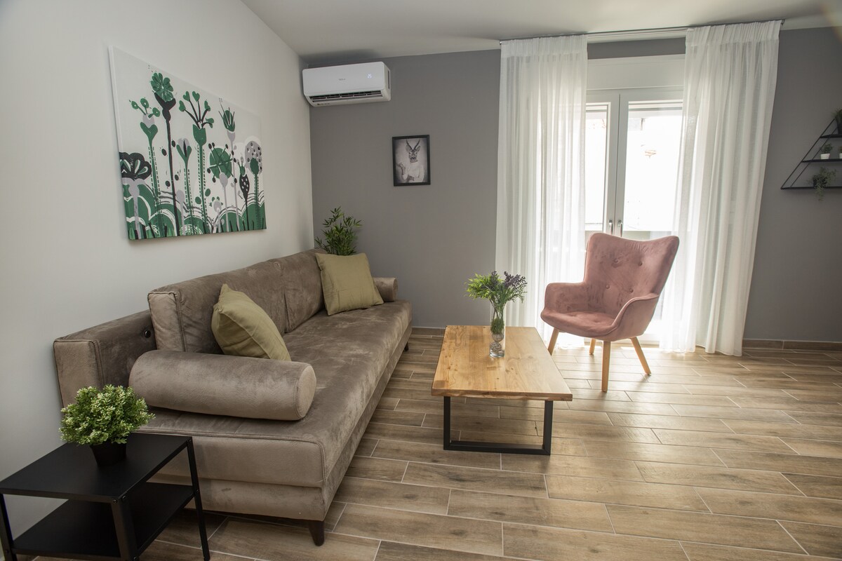 A comfortable living area is presented, featuring a neutral-colored sofa adorned with decorative pillows. A wooden coffee table is centrally placed, complemented by a vase of fresh flowers. A pink accent chair adds a pop of color, with natural light filtering through sheer curtains.