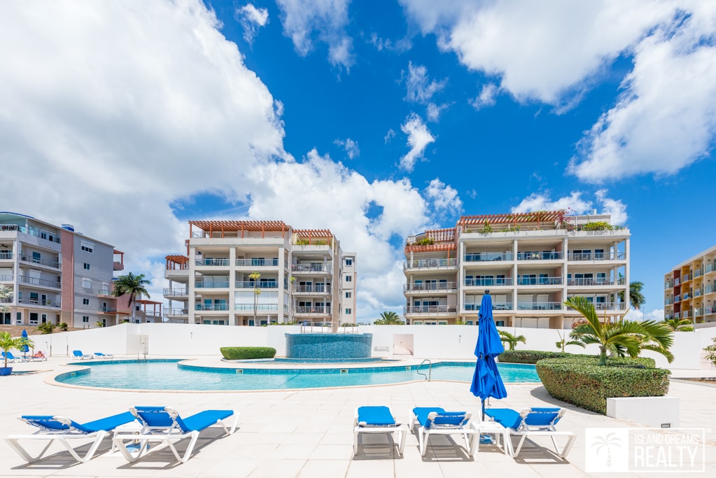 A spacious outdoor area features a circular swimming pool surrounded by a sun deck. Blue lounge chairs are arranged around the pool, while a cascading water feature adds a serene touch. Bright clouds scatter across the sky above the villa buildings in the background.