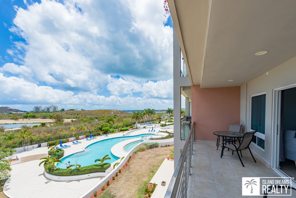 A spacious balcony offers a view of a lagoon-style pool surrounded by lounge chairs and lush landscaping. The cloudy sky reflects tranquility, while the nearby greenery enhances the sense of privacy. A small table and chairs are set for outdoor relaxation.