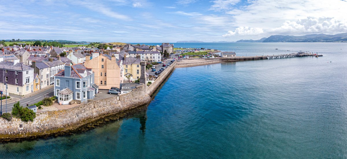 A panoramic view of a coastal town captures colorful buildings lining the water's edge. A pier extends into the calm sea, while distant mountains create a scenic backdrop beneath a partly cloudy sky. The image conveys a serene atmosphere, inviting exploration of the area.