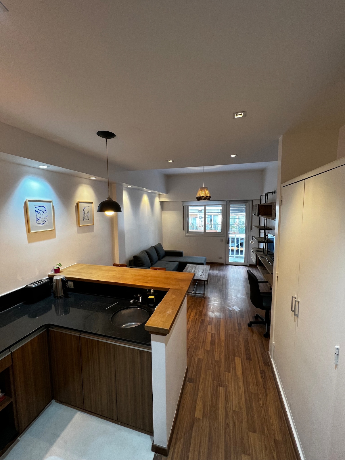 An open-concept living area features a modern kitchen with wooden countertops and sleek cabinetry. A comfortable seating area is arranged around a coffee table, with large windows allowing natural light to fill the space. Light fixtures are strategically placed, enhancing the contemporary design.