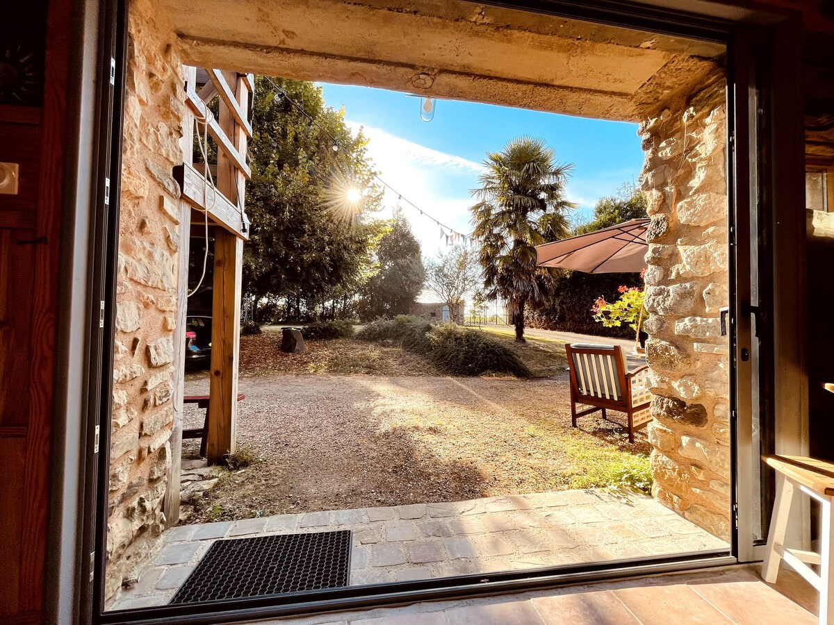 The doorway frames a view of the garden and surrounding trees, illuminated by soft sunlight. A pathway leads into the outdoor space, where seating can be seen. A patio umbrella provides shade, while decorative lights hang in the background.