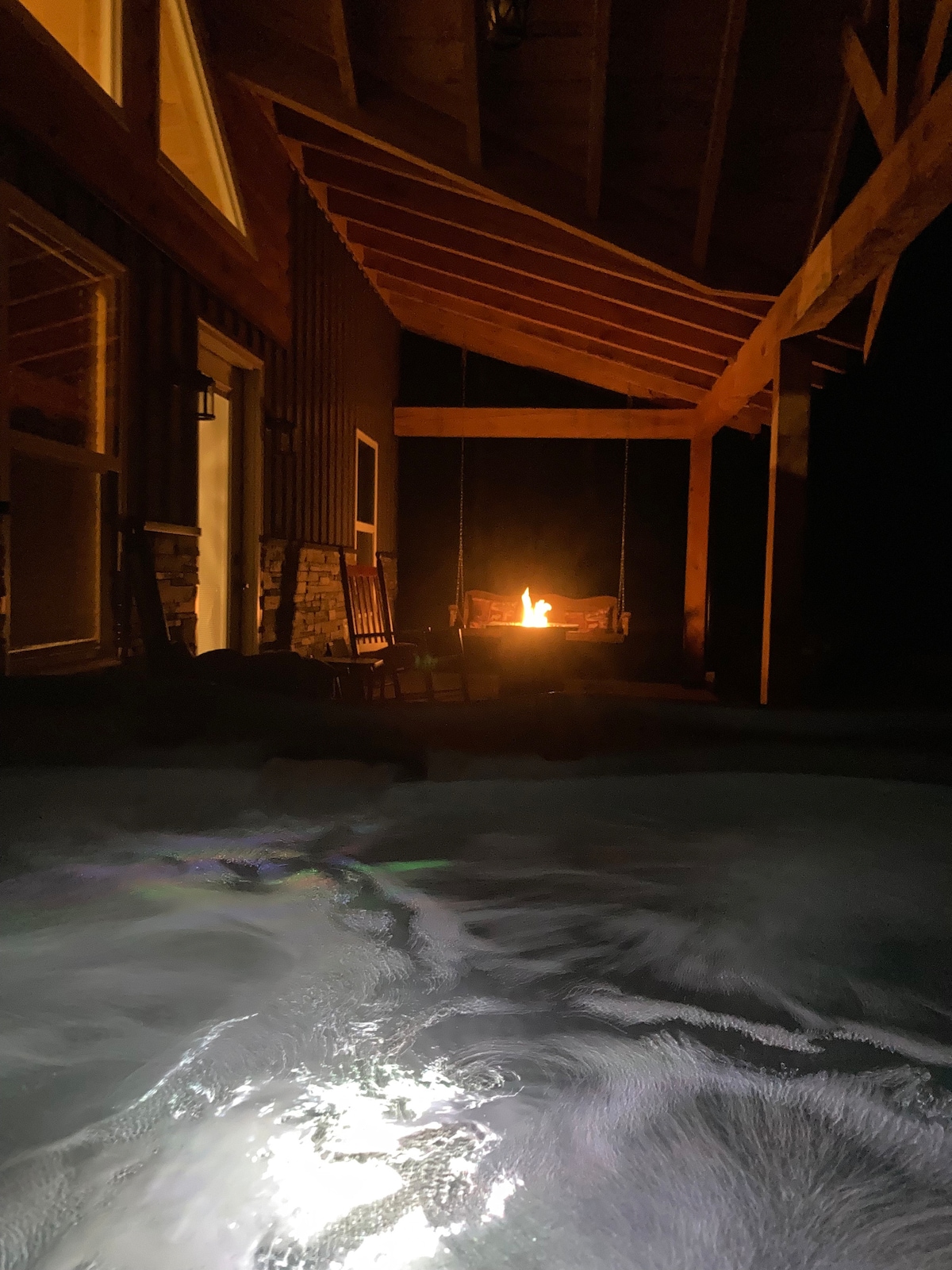 The image captures a serene outdoor space illuminated by a fire pit. Water gently ripples in the foreground, indicating a hot tub. Wooden rocking chairs are positioned nearby, creating a cozy atmosphere under the wooden structure's overhang.