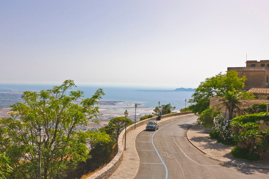 A scenic view is presented from a winding road overlooking the coastline. Lush trees frame the image, while a distant horizon reveals the sea and surrounding landscape. The road is paved and curves gently, leading to nearby buildings and greenery.
