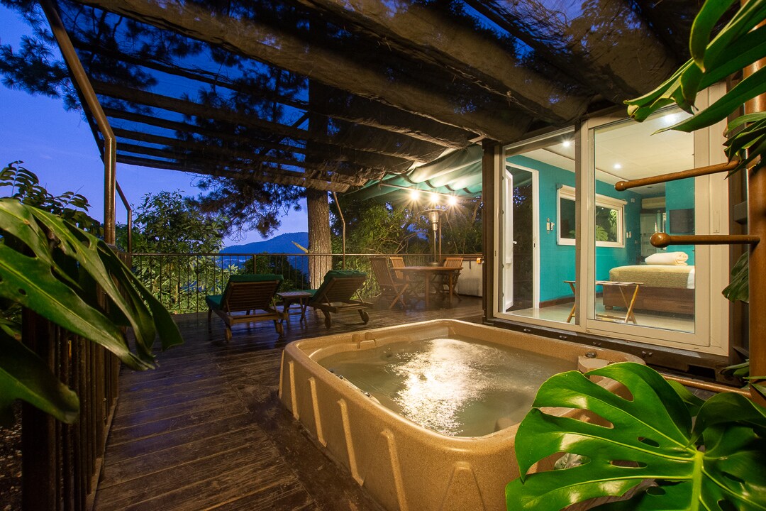 A private deck is surrounded by lush greenery, featuring a relaxing jacuzzi and two lounge chairs facing the stunning views. Soft lighting enhances the inviting ambiance, while the room inside is visible through the expansive glass doors that connect the indoor and outdoor spaces.