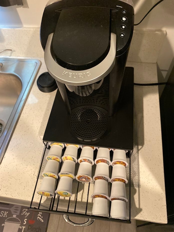 A modern coffee maker is placed on a countertop beside a sink, featuring a drawer filled with various coffee pods. The machine is designed for single-serve brewing and is equipped with an accessible control panel.
