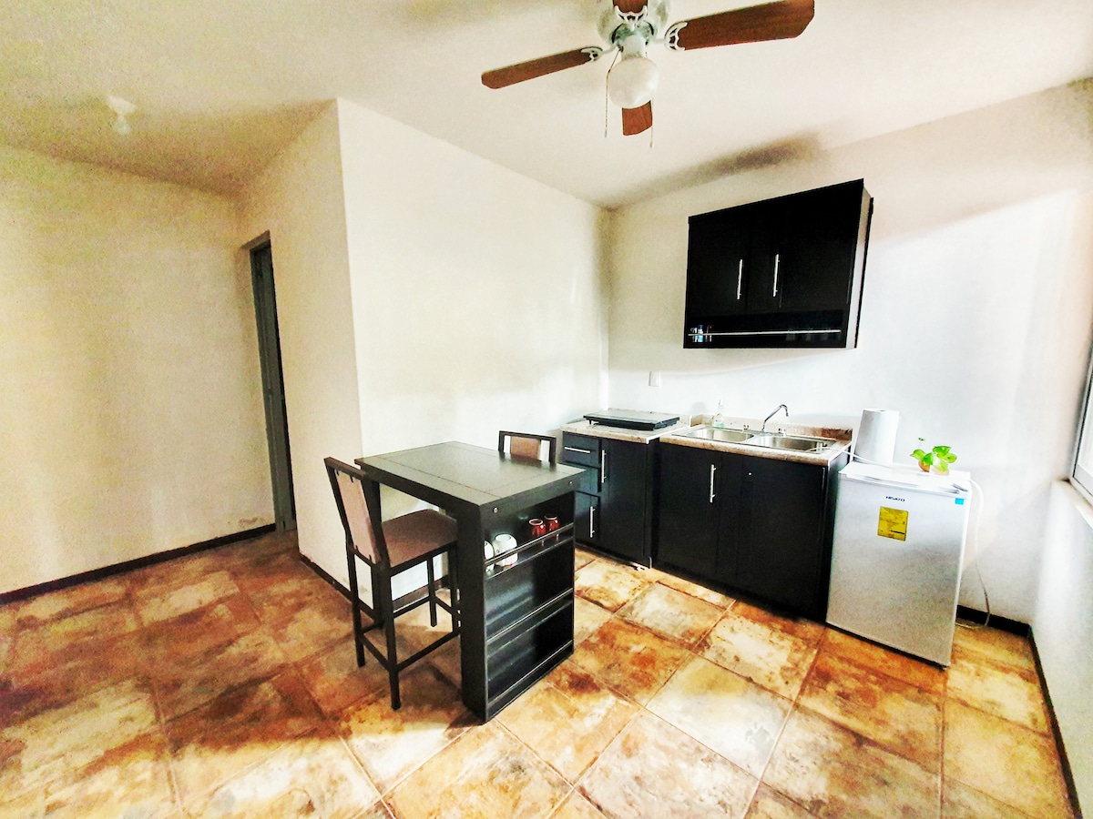 A compact kitchen area features black cabinetry and a sink. A small table with two chairs is positioned nearby. An electric stove and mini refrigerator are also present, with tile flooring providing a neutral backdrop. Natural light enters through a nearby window.
