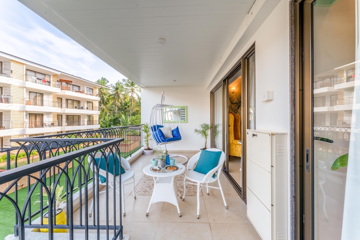 A spacious balcony features a blend of indoor and outdoor seating, including a hanging chair and a small dining table. Lush greenery is visible in the background, with light streaming in through the glass doors that connect the balcony to the living area.