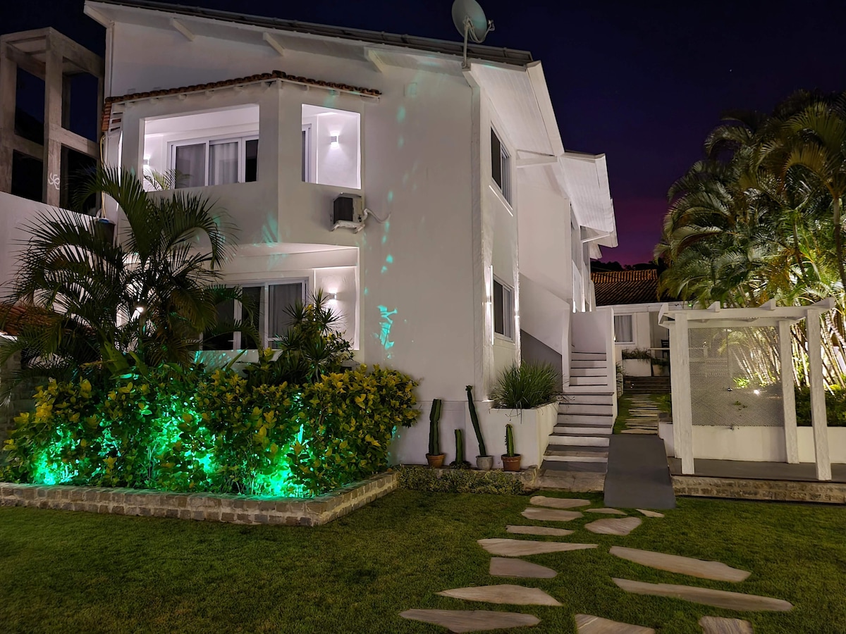The two-story white building is set against a twilight sky, illuminated by soft green lights. A pathway of stone leads through a well-kept garden, featuring diverse plants and shrubs. The structure showcases large windows and a welcoming entrance, enhancing the modern design.