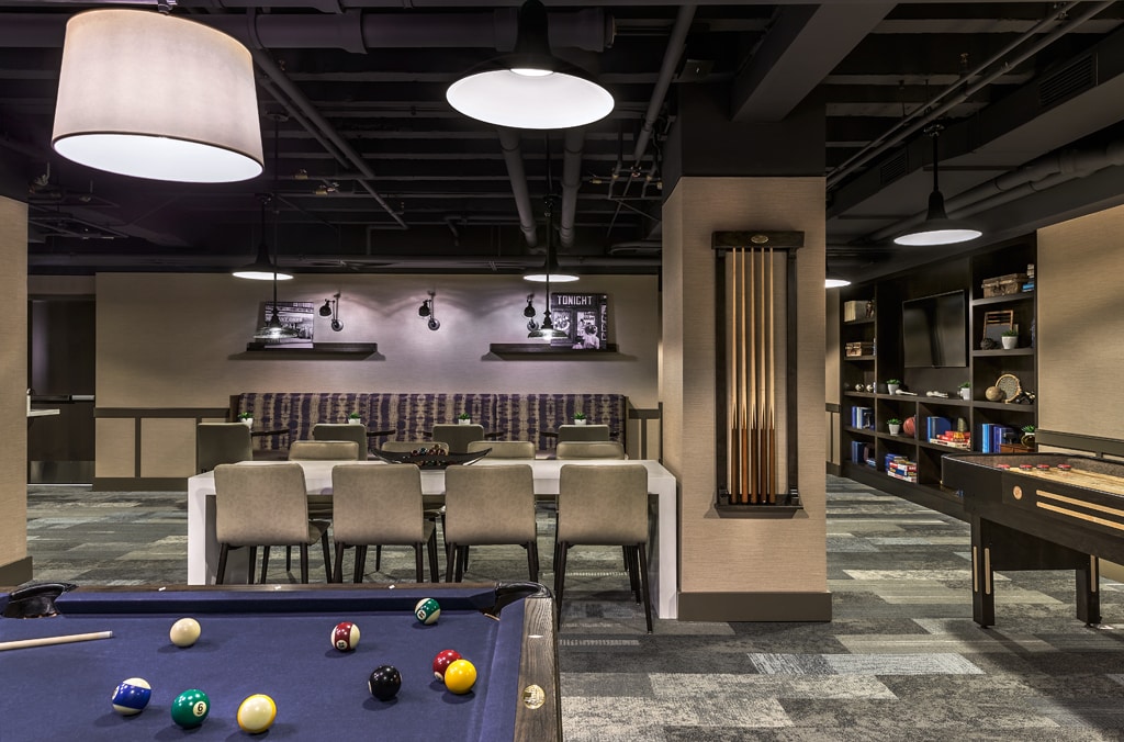 A recreational area features a billiards table with colorful balls scattered across the felt. Surrounding seating includes several modern chairs at a long table, with shelves displaying games and books in the background. Warm lighting from overhead fixtures enhances the inviting space.