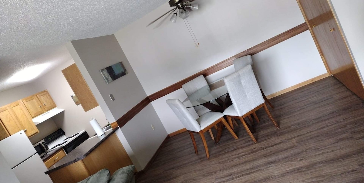 A spacious dining area is visible, featuring a glass table surrounded by four light-colored chairs. A ceiling fan hangs above, while the kitchen area can be seen in the background, showcasing wooden cabinetry and modern appliances. Natural light fills the room through a nearby window.