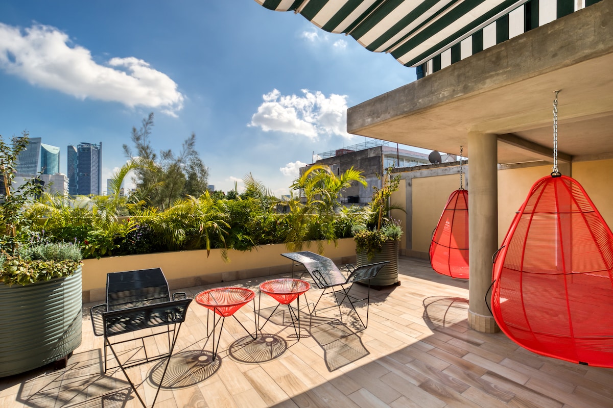 An inviting roof garden features spacious seating areas, including four metal chairs and two hanging red chairs. Lush greenery creates a serene environment, with views of urban buildings in the distance under a clear blue sky, complemented by a striped awning for shade.