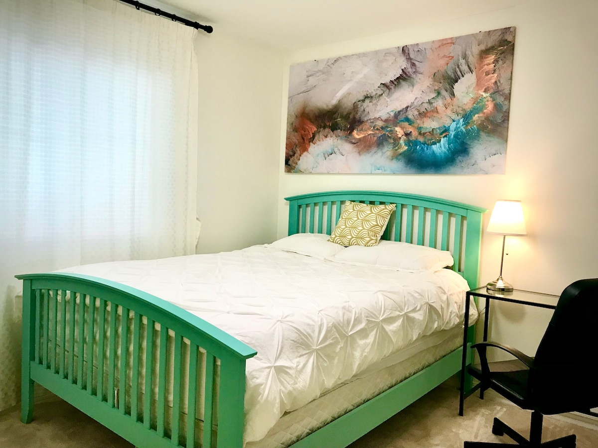 A spacious bed with a turquoise frame is centered in the room, complemented by white linens. A decorative pillow adds a touch of design. A vibrant abstract artwork hangs above the bed, while a lamp and a black chair are positioned nearby.