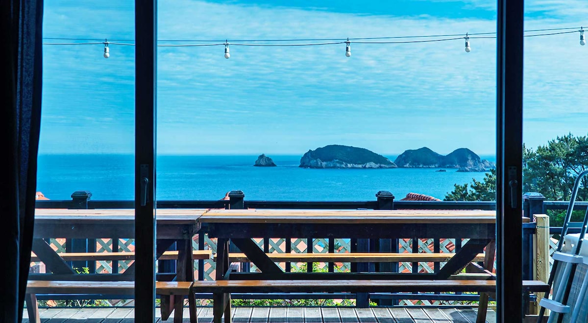 A panoramic view of the ocean is framed by large glass doors. The tranquil water reflects the sky, while distant islands are visible on the horizon. A spacious wooden deck is seen, furnished with a long bench, inviting relaxation and enjoyment of the scenery.
