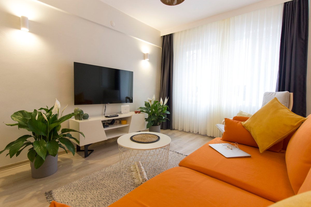 A comfortable living area is presented with an orange sectional sofa, complemented by neutral-colored cushions. A round coffee table sits at the center, surrounded by potted plants. Natural light filters through sheer curtains, creating a bright and inviting space.