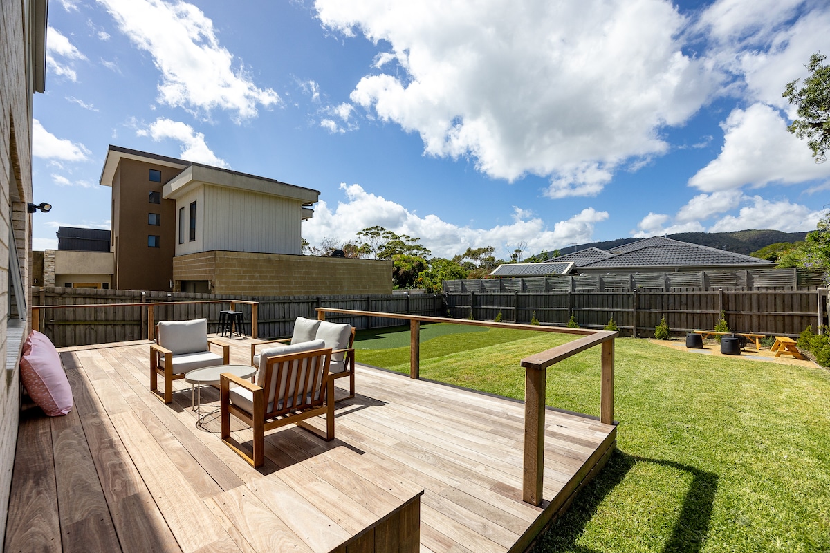 A spacious backyard is displayed, featuring a wooden deck with seating for four. The well-maintained grass area offers ample space for outdoor activities, while a fence provides privacy. A variety of trees and shrubs are present in the background, enhancing the outdoor setting.