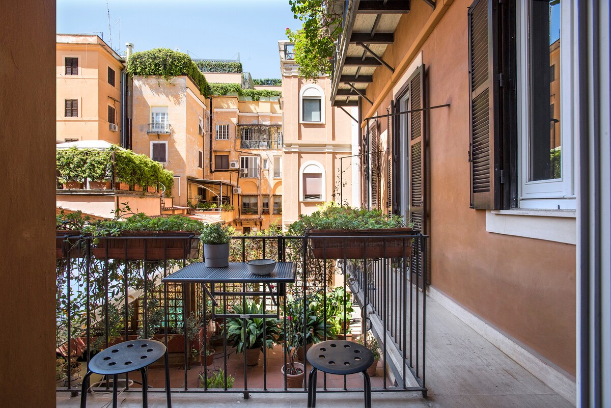 A small balcony is shown with a table and stools surrounded by potted plants. The view features terracotta buildings with greenery, creating a serene outdoor space perfect for relaxation. Natural light illuminates the area, contributing to a welcoming atmosphere.